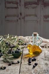 Black olives and jug of oil placed near fresh tree twigs on wooden table in rustic kitchen - ADSF37550