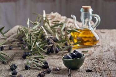 Black olives and jug of oil placed near fresh tree twigs on wooden table in rustic kitchen - ADSF37552