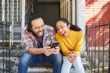 Young happy unshaven Mexican man with cellphone interacting with laughing female beloved while sitting on staircase in sunlight - ADSF37576