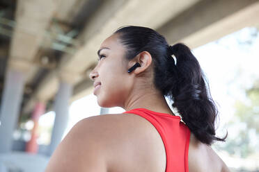 Positive Hispanic female athlete in sportswear listening to music in earphones after workout against metal net - ADSF37579