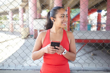 Positive Hispanic female athlete in sportswear listening to music in earphones while using mobile phone after workout against metal net - ADSF37581