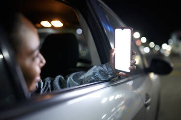 Seitenansicht einer lächelnden weiblichen Passagierin auf dem Rücksitz, die in einer nächtlichen Stadt auf ihrem Mobiltelefon mit leerem Bildschirm surft - ADSF37587