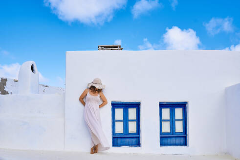 Unerkennbare Frau in Sommerkleidung, die ihr Gesicht mit einem Hut bedeckt, während sie vor einem ländlichen weißen Haus mit blauen Fensterläden in einem sonnigen Land steht - ADSF37593