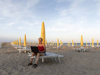 Glückliche ältere Geschäftsfrau sitzt mit Laptop am Strand - FLLF00674