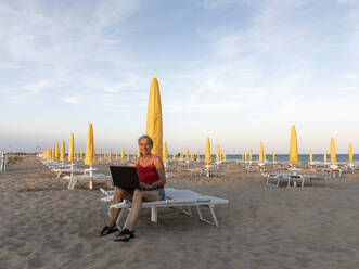 Happy senior businesswoman sitting with laptop at beach - FLLF00674