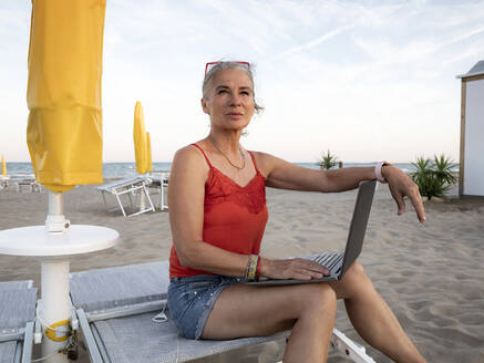 Nachdenkliche ältere Geschäftsfrau sitzt mit Laptop am Strand - FLLF00675