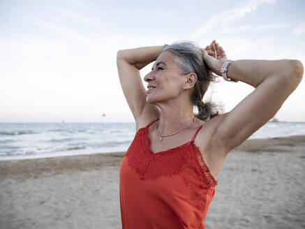 Senior woman with hands behind head standing at beach - FLLF00684