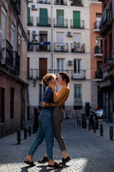 Ganzkörperfrauen in Freizeitkleidung, die sich umarmen und lachen, während sie bei einem romantischen Date an einem sonnigen Tag in der Stadt auf dem Bürgersteig stehen - ADSF37603