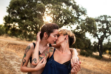 Tattooed girlfriends with closed eyes and touching each other while hugging and kissing in nature in cloudy day - ADSF37606