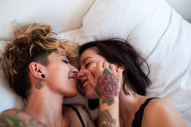 Fröhliche tätowierte Frauen in Unterwäsche, die sich gegenseitig anschauen und lachen, während sie auf einem bequemen Bett zu Hause zusammen ausruhen - ADSF37611