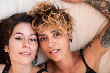 Fröhliche tätowierte Frauen in Unterwäsche, die lachend in die Kamera schauen, während sie gemeinsam auf einem bequemen Bett zu Hause ruhen - ADSF37616