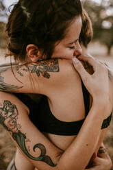 Tattooed girlfriends with closed eyes and touching each other while hugging in nature in cloudy day - ADSF37619