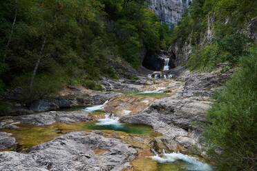 Kaskade fließt von Steinstufen in den plätschernden Fluss inmitten hoher grüner Bäume an einem Sommertag in der Natur Spaniens - ADSF37645