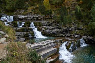 Schnelle Kaskade, die von einer Steintreppe in einen plätschernden Fluss inmitten hoher grüner Bäume an einem Sommertag in der Natur Spaniens fließt - ADSF37646