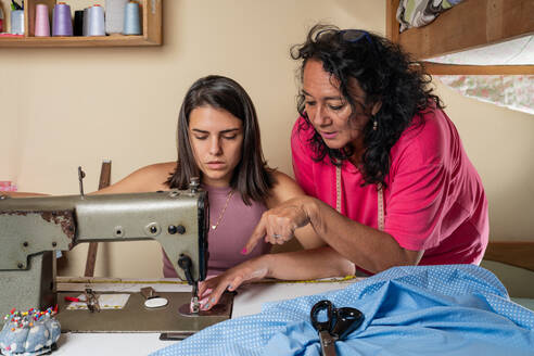 Spanische Näherin, die ihrer Tochter das Nähen an einer professionellen Maschine beibringt, während sie an einem Tisch mit Stoffen und verschiedenen Materialien in einer hellen Werkstatt arbeitet - ADSF37652