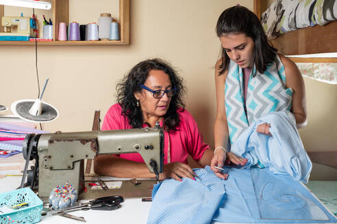 Spanische Näherin mit Tochter am Tisch mit Nähmaschine und verschiedenen Textilien bei der Arbeit in der Werkstatt - ADSF37653