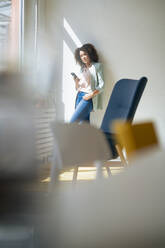 Mature businesswoman with smart phone leaning on wall at office - JOSEF13017