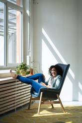 Fröhliche Geschäftsfrau sitzt auf einem Stuhl am Arbeitsplatz - JOSEF13033