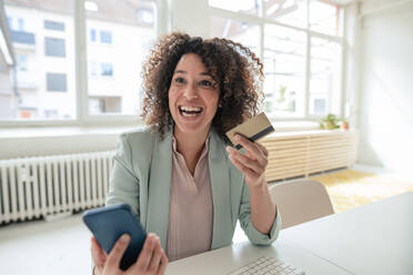 Fröhliche Geschäftsfrau mit Kreditkarte und Smartphone am Arbeitsplatz - JOSEF13061