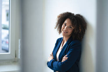 Smiling businesswoman with curly hair leaning on wall - JOSEF13076
