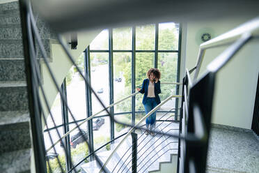 Businesswoman talking through mobile phone standing on staircase in office - JOSEF13097