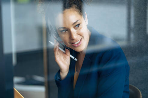 Businesswoman talking through headset sitting in soundproof cabin at office - JOSEF13117