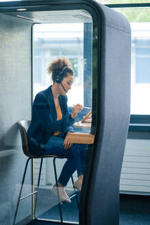 Mature businesswoman with headset using tablet PC sitting in soundproof cabin at workplace - JOSEF13122