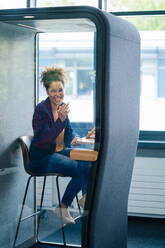 Cheerful businesswoman sitting in soundproof cabin at office - JOSEF13123