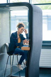 Mature businesswoman sitting in soundproof cabin at workplace - JOSEF13124