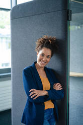 Smiling businesswoman with arms crossed leaning on soundproof cabin at office - JOSEF13133