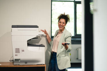Cheerful businesswoman standing by computer printer at workplace - JOSEF13150