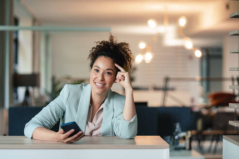 Lächelnde Geschäftsfrau mit Mobiltelefon am Schreibtisch im Büro - JOSEF13151