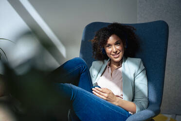 Smiling businesswoman with smart phone sitting on chair at office - JOSEF13177