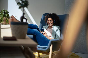 Happy businesswoman using mobile phone sitting on chair at workplace - JOSEF13180