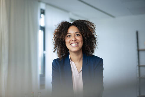 Happy businesswoman with curly hair clenching teeth at workplace - JOSEF13204