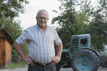 Senior farmer with hands on hips standing in front of tractor - OSF00906