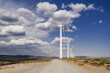 Empty road by wind turbines lined up on sunny day - EBBF06286