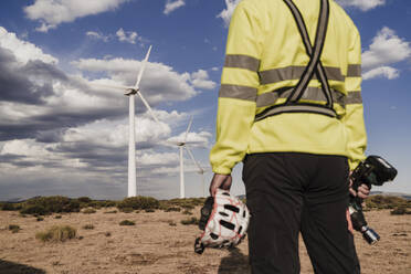 Technician holding work tool and helmet standing at wind farm - EBBF06295