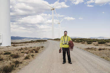 Engineer with helmet and rope standing on road at wind farm - EBBF06298