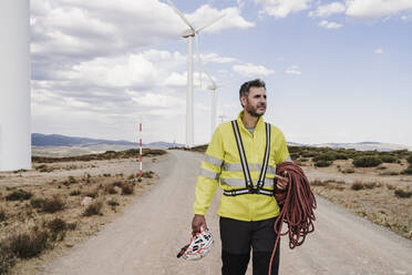 Technician with rope and helmet walking on road at wind farm - EBBF06299