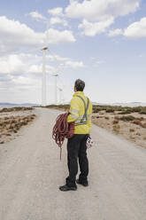 Technician standing on road looking at wind turbines at wind farm - EBBF06303