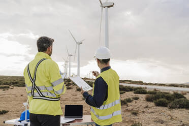 Engineer discussing with colleague looking at wind turbines at wind farm - EBBF06310