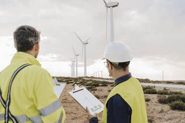 Technicians holding clipboards looking at wind turbines at wind farm - EBBF06311