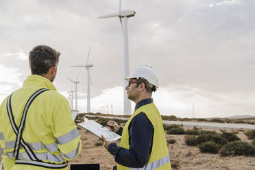 Engineer holding clipboard looking at colleague at wind farm - EBBF06312