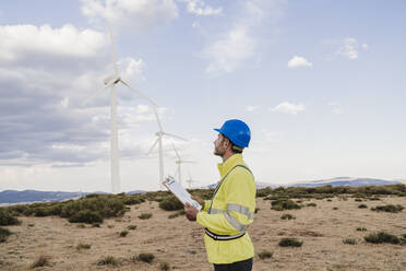 Engineer holding clipboard looking at wind turbine - EBBF06319