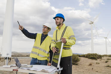 Engineer explaining and talking to colleague holding solar panel at wind farm - EBBF06324