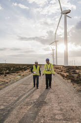 Technician and colleague discussing walking on dirt road at wind turbines - EBBF06339