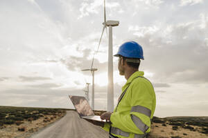 Älterer Ingenieur mit Laptop, der sich die Turbinen in einem Windpark ansieht - EBBF06342