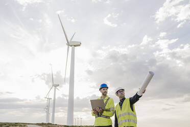 Engineer with blueprint discussing with colleague holding laptop at wind farm - EBBF06344