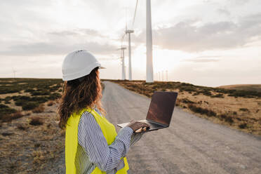 Ingenieur mit Laptop betrachtet Windturbinen in einem Bauernhof bei Sonnenuntergang - EBBF06357